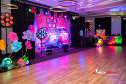 Wieczór tematyczny Flower Power - rzut na dekorację główną sali: duża grafika tematyczna w formie backdropu, kolorowe kwiaty XXL, parawan DJ-a z barwną tematyczną grafiką, zestaw techniki.